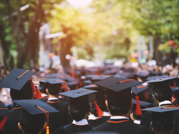 A number of college graduates dressed in traditional cap and gown attire, walking in a group outdoors.