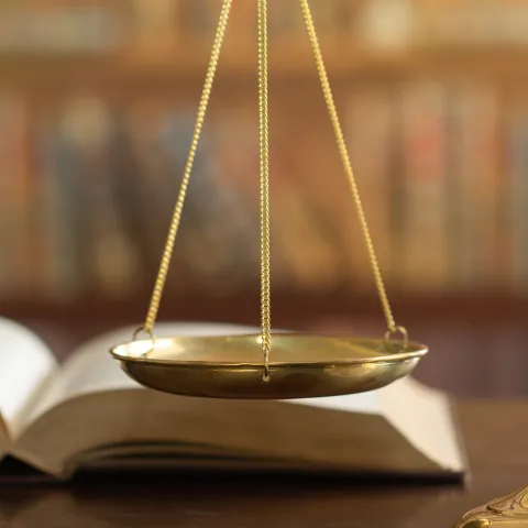 A close up of an open book on a wood table, with a bronze scales of justice and a wooden gavel visible in the foreground.